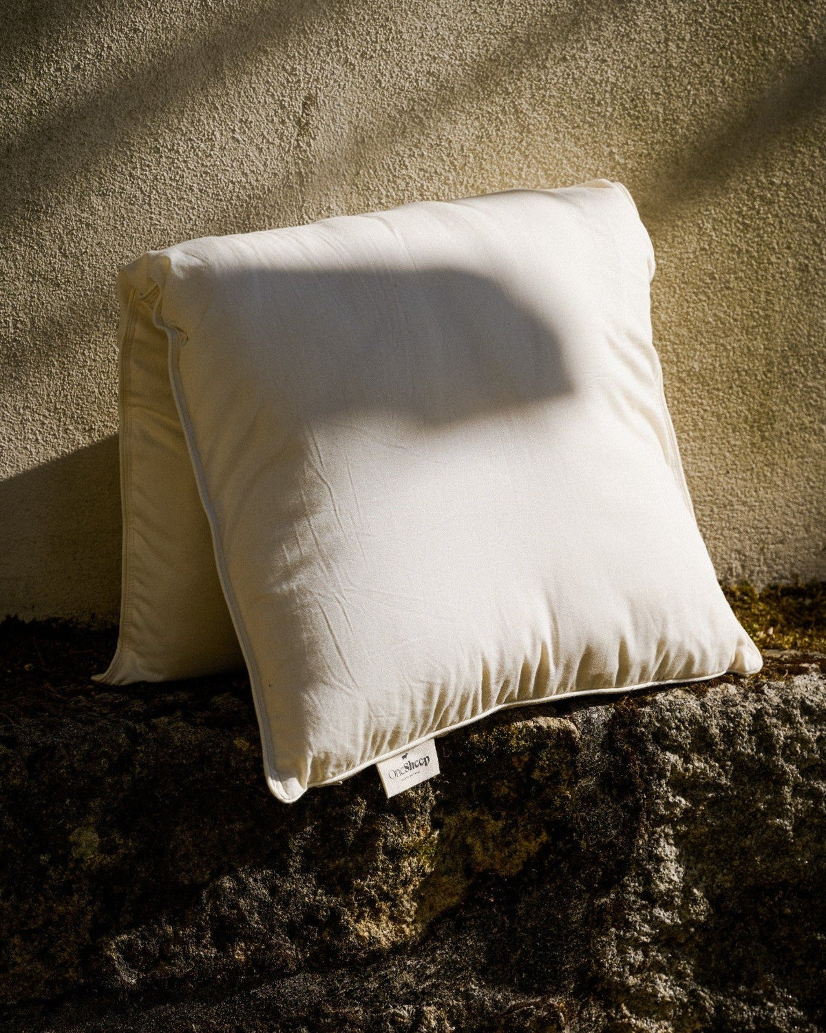 OneSheep Responsible Down Pillow in natural white organic cotton cover, leaning against a stone wall in warm natural light.